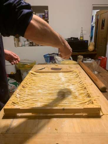 A person is making homemade pasta, spreading and dusting flour over freshly cut noodles on a wooden table in a cozy kitchen with various utensils nearby.