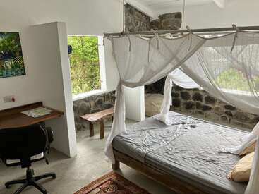 L'image représente une chambre sereine avec un lit à baldaquin, un bureau et une chaise, avec un mur en pierre et une fenêtre offrant une vue sur la verdure luxuriante à l'extérieur.