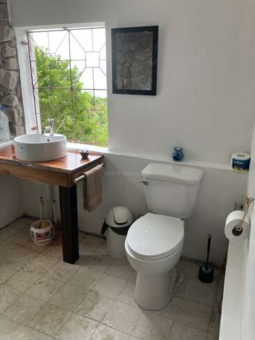 L'image représente une salle de bains avec des toilettes, un lavabo et une fenêtre blancs, avec une table en bois, un mur en pierre et un sol en carrelage, le tout sur un fond naturel serein.