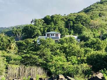 L'image représente une maison blanche au toit vert, nichée au milieu d'arbres et de feuillages luxuriants sur le flanc d'une colline, sur fond de ciel gris.
