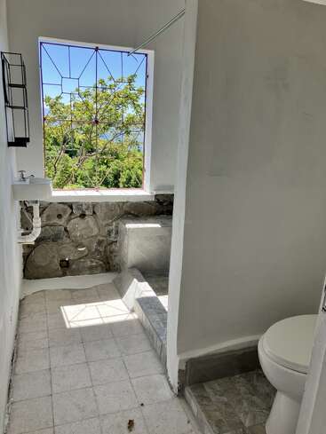 L'image représente une petite salle de bains avec une fenêtre donnant sur des arbres et un ciel bleu, avec des toilettes, un lavabo, un mur en pierre et un sol carrelé blanc.