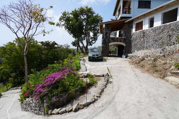 L'image représente une scène sereine d'une maison avec une grande cour, comprenant une allée et une pelouse bien entretenue, entourée d'arbres et de buissons, dans une atmosphère claire et ouverte.