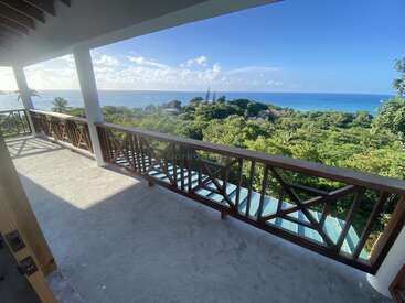 L'image représente un balcon serein avec une balustrade en bois, surplombant un paysage verdoyant et l'océan, dans un ciel bleu vif avec des nuages blancs.