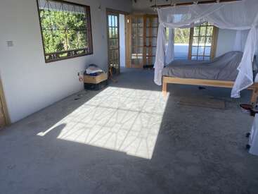L'image représente une chambre sereine avec un lit, des rideaux blancs et un sol en béton, baignée par la lumière du soleil qui traverse une fenêtre et une porte.