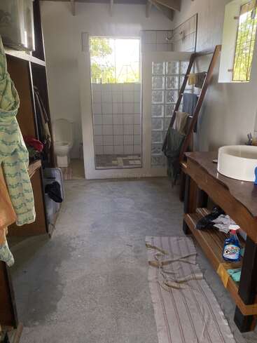 L'image représente une salle de bain avec des toilettes, une douche et un lavabo, avec une table en bois, une échelle et divers objets éparpillés sur le sol et les étagères.