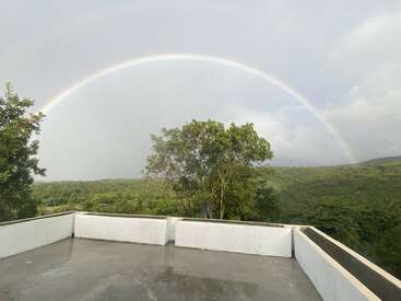 L'image représente une scène sereine d'arc-en-ciel s'étendant sur un paysage verdoyant, avec un toit en béton et des arbres au premier plan.