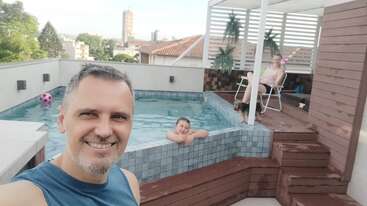 Un hombre se hace un selfi junto a una pequeña piscina en la azotea, sonriendo. Hay un niño en el agua y una mujer descansa en una tumbona cerca de allí.