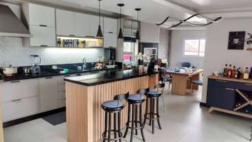 Una cocina moderna con una isla con encimera negra, cuatro taburetes de bar, lámparas colgantes, armarios blancos, electrodomésticos de cocina y un comedor con un espacio habilitado como oficina en casa.