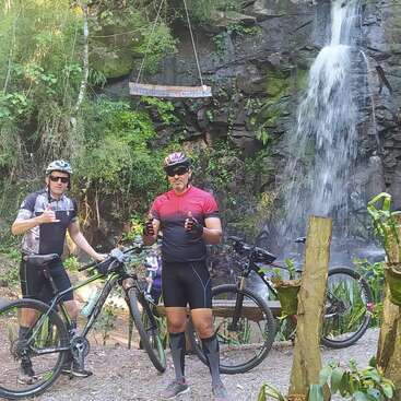 Zwei Radfahrer in Radbekleidung posieren mit ihren Mountainbikes vor einem üppig grünen Wasserfall, umgeben von Felsen und Grün, und sehen glücklich und entspannt aus.