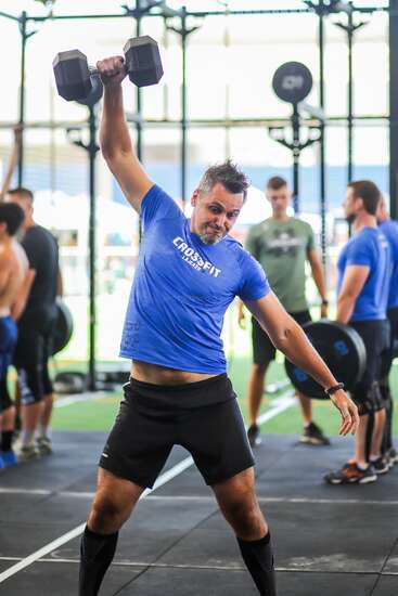 Ein Mann in einem blauen „CrossFit“-Shirt hebt in einem Fitnessstudio eine Hantel über den Kopf, während im Hintergrund andere trainieren und Gewichte stemmen. Es herrscht eine energiegeladene Atmosphäre.