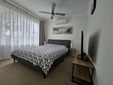 Un acogedor dormitorio con una gran ventana, colcha con motivos de corazones, TV sobre un soporte de madera, mesillas de noche, ventilador de techo y una tranquila obra de arte playera sobre la cama.