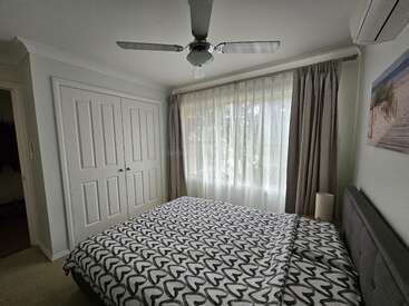 Este acogedor dormitorio cuenta con una cama doble con edredón estampado, una gran ventana con cortinas transparentes, ventilador de techo, armario, aire acondicionado, mesilla de noche y arte mural.