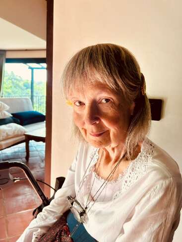 An elderly woman with gray hair, wearing a white blouse and glasses, smiling gently while sitting indoors. Sunlight gently illuminates her face, creating a warm atmosphere.
