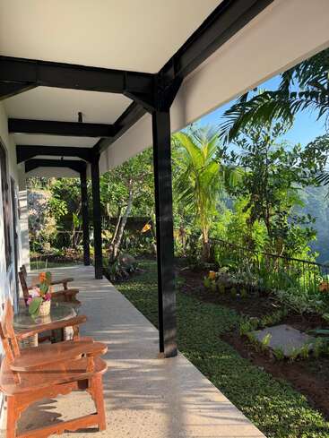 A peaceful covered patio with wooden chairs and a glass table overlooks a lush, green garden with tropical plants, basking in bright, natural sunlight.