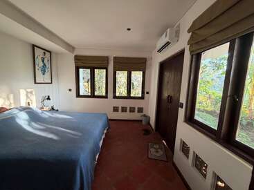 A cozy bedroom with a blue bedspread, three large windows, green roman blinds, potted plants outside, artwork, bedside lamp, air conditioner, and terracotta floor.