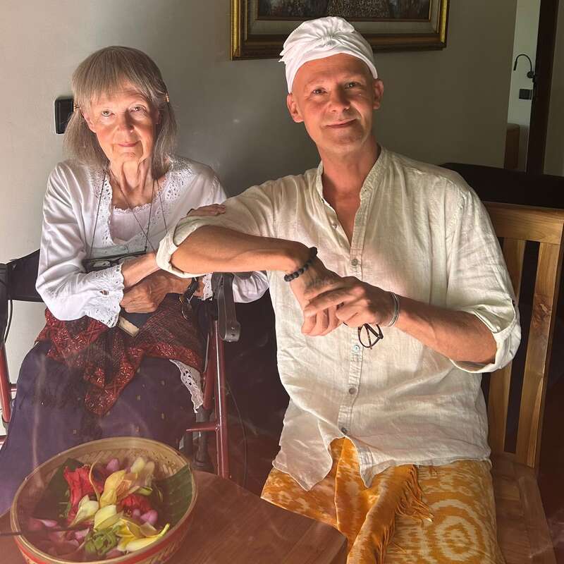 An elderly woman and a man sit together indoors, both dressed in light traditional clothing. The man wears a white headscarf. A colorful fruit bowl is present.
