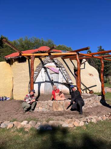 Three people pose in front of a unique, dome-shaped adobe building with wooden beams, stone landscaping, and bright blue skies, enjoying a sunny day outdoors together.