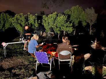 Three people sit around a glowing campfire at night, surrounded by trees. Two are seated, one stands nearby. A table holds drinks, creating a cozy atmosphere.