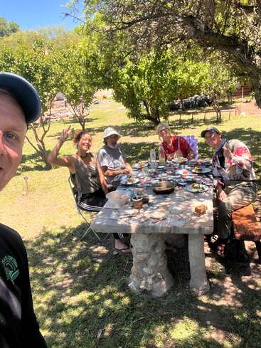 Five people are enjoying a meal outdoors at a stone table under a tree, smiling and making peace signs. The sunny setting appears relaxed and cheerful.