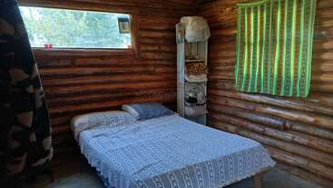 This cozy log cabin bedroom has a simple bed with patterned bedding, a small shelving unit with blankets and clothes, and windows providing natural light and forest views.