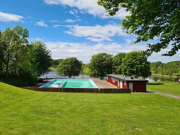 Une piscine extérieure sereine est entourée d'une herbe verte luxuriante, d'arbres vibrants et d'un ciel bleu clair, à côté d'un lac paisible et réfléchissant et d'un bâtiment rouge.