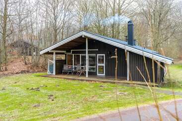 Une petite cabane en bois sombre avec un toit en pente se trouve dans une clairière herbeuse. Des meubles d'extérieur sont posés sur le porche, entourés d'arbres nus et d'une atmosphère paisible.
