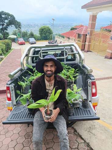 Un homme coiffé d'un chapeau est assis sur le hayon ouvert d'une camionnette, tenant un pot de fleurs, entouré d'autres plantes, souriant dans un cadre extérieur pittoresque.