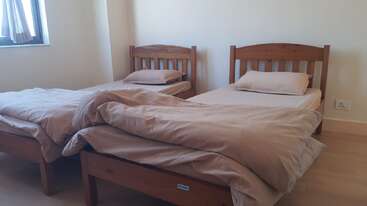 Cette image montre une chambre simple et bien rangée avec deux lits simples en bois, une literie beige, des murs clairs, un sol en bois et une fenêtre laissant entrer la lumière naturelle.