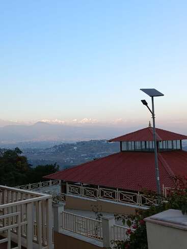 Une vue panoramique mettant en scène un bâtiment au toit rouge, une balustrade, un lampadaire solaire, des arbres verts, des collines lointaines et des montagnes enneigées sous un ciel bleu limpide.