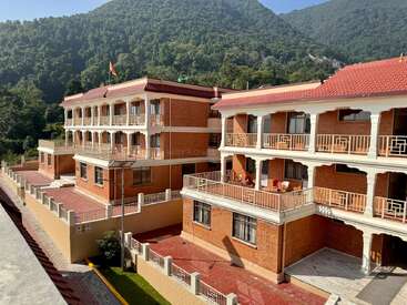 Un magnifique complexe immobilier en briques, avec balcons et toits rouges, se dresse au milieu de collines verdoyantes sous un ciel bleu limpide, dégageant une atmosphère paisible et un charme naturel.
