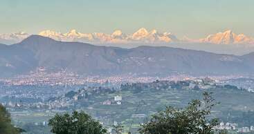 Cette image montre un paysage pittoresque avec une ville au premier plan, des collines verdoyantes et de majestueuses montagnes enneigées à l'arrière-plan, sous un ciel clair.
