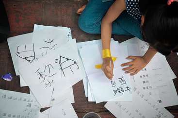 The image depicts a child sitting on the floor, surrounded by papers with Chinese characters, engaged in writing with a brush.