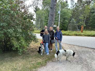 Quatre personnes se tiennent au bord d'une route rurale avec deux chiens noirs et blancs en laisse. Un feuillage vert dense et de grands arbres entourent ce cadre extérieur paisible.