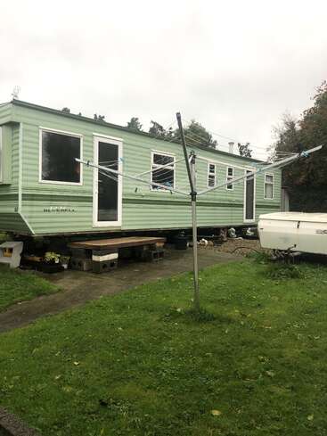 The image depicts a light green mobile home with white trim, featuring a clothesline in the foreground and a lush grassy area in front of it.