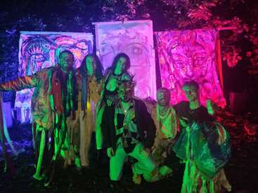 Six people in colorful costumes pose outdoors at night, illuminated by green and pink lights. Behind them are three large, vibrant, surreal paintings with faces.