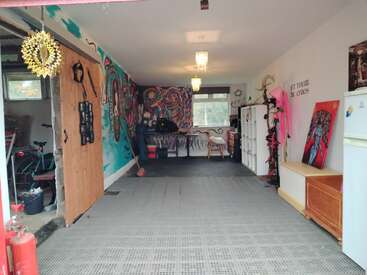 A colorful, eclectic room with vibrant murals, artwork, and creative decorations. There are workspaces, wooden furniture, a bicycle, fridge, and the phrase "Let there be chaos."