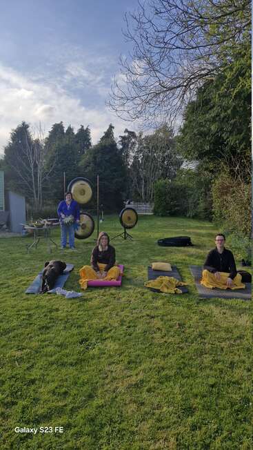 Three people and a dog sit on yoga mats outside, surrounded by green grass and trees. Large gongs and bowls suggest a sound healing or meditation session.