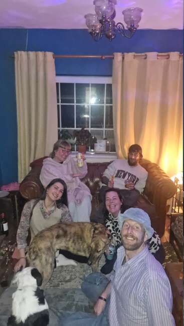 Cinco amigos se reúnen en una acogedora sala de estar, posando alegremente con bebidas y aperitivos, acompañados por dos perros, creando juntos un ambiente cálido, alegre y relajado.
