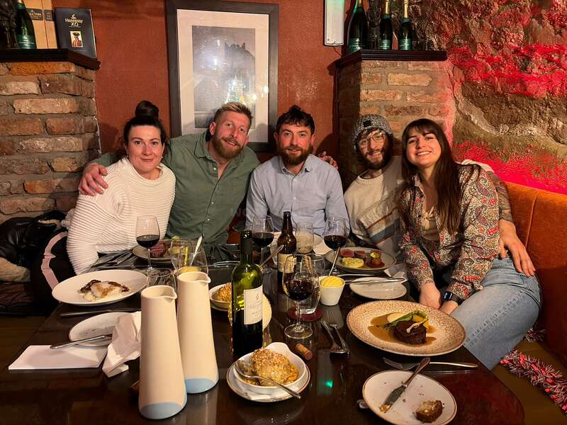 Five friends sit closely together at a cozy restaurant, smiling and enjoying dinner. The table is filled with food, drinks, and a warm, inviting atmosphere.