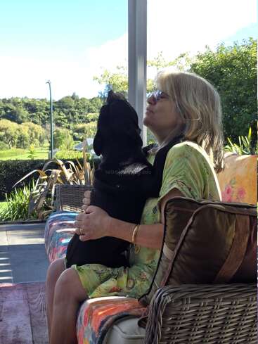 Uma mulher de óculos escuros está sentada em um sofá colorido ao ar livre, abraçando um cachorro preto. Eles estão rodeados de vegetação e aproveitando juntos um dia tranquilo e ensolarado.