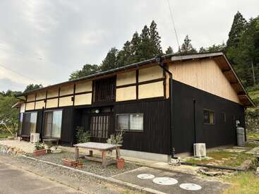 Das ist ein traditionelles japanisches Haus mit schwarzen Holzwänden, Glasschiebetüren, einem schrägen Dach und Pflanzen im Außenbereich, umgeben von Bäumen unter einem bewölkten Himmel.