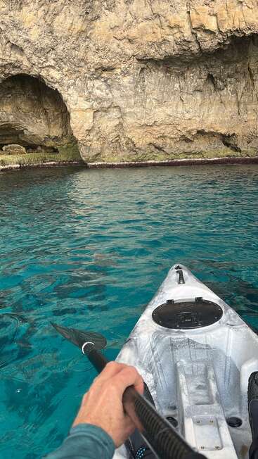 A person in a kayak paddles through clear turquoise water towards a rocky cliff with a cave, surrounded by textured stone and peaceful, vibrant scenery.