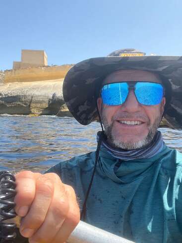 Un hombre con un sombrero de ala ancha, gafas de sol reflectantes y una camisa azul sonríe mientras navega en kayak cerca de la costa rocosa bajo un cielo azul despejado.