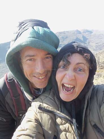 A cheerful man and woman, both wearing hooded jackets, take a close-up selfie outdoors with mountains in the background, enjoying a fun, energetic moment together.