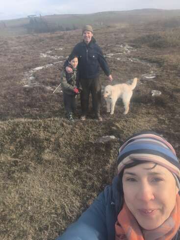 Une femme prend un selfie en plein air. Derrière elle, un homme, un enfant et un chien blanc au pelage duveteux se tiennent côte à côte dans un paysage ouvert et verdoyant, sous un ciel nuageux.