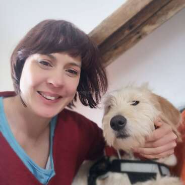 Una mujer sonriente, con el pelo castaño corto, se hace un selfi mientras sostiene con delicadeza a un perro peludo y de pelaje claro. Ambos parecen felices, compartiendo un momento acogedor y alegre en el interior.
