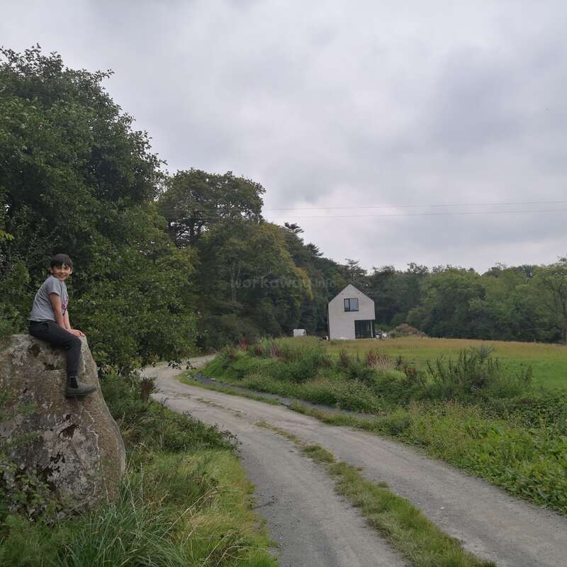 Uma criança está sentada em uma grande pedra ao lado de uma estrada rural sinuosa, com uma vegetação exuberante, céu nublado e uma casa branca cercada por campos ao longe.