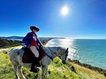 Das Bild zeigt eine Person, die auf einem Pferd auf einer grasbewachsenen Klippe mit Blick auf ein Gewässer reitet, während die Sonne hell in den Himmel scheint.