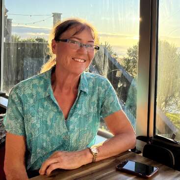 Una mujer con gafas, vestida con una camisa azul estampada, sonríe sentada a una mesa cerca de una ventana durante la puesta de sol. A su lado hay un teléfono.