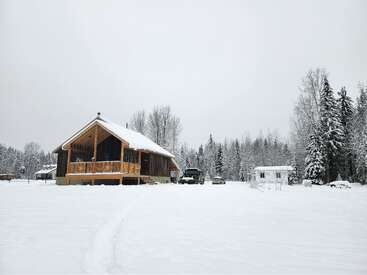 A cozy wooden cabin sits in a snowy landscape, surrounded by tall trees. Vehicles and a small shed are nearby, creating a peaceful winter retreat scene.